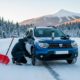 Hombre colocando cadenas de nieve en un Dacia Duster azul en una carretera de montaña nevada, con una pala roja y un termo al lado.