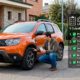 Un hombre revisa la presión del neumático de un Dacia Duster naranja estacionado en la calle. A la derecha, una infografía superpuesta muestra un checklist con el título '¿GASTA MÁS GASOLINA DE LO NORMAL? 5 Culpables No del Motor', enumerando factores como la presión de neumáticos, carga excesiva, aerodinámica, mantenimiento y estilo de conducción.